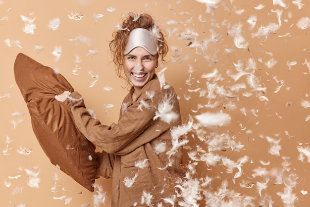 Positieve blije jonge vrouw heeft kussengevecht na het ontwaken heeft plezier en lacht gekleed in comfortabele pyjama blinddoek op voorhoofd vormt binnenveren die rondvliegen slaaprusthuisconcept