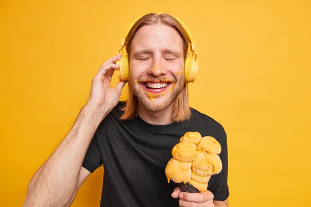 Positieve blije roodharige man sluit ogen en glimlacht gelukkig geniet van favoriete liedje houdt hand op koptelefoon houdt groot lekker ijs heeft vrije tijd tijdens vrije dag in de zomer geïsoleerd op gele muur