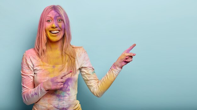 positieve blonde vrouw vuil met kleurrijke poeder, iets op lege ruimte adverteert, lacht vrolijk, geniet van Indiase fest, geïsoleerd tegen blauwe muur. Promotie concept