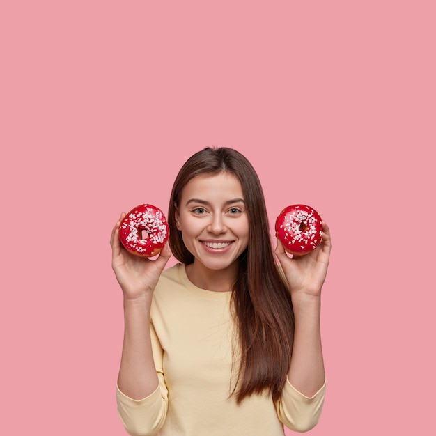 Positieve brunette vrouw draagt twee donuts in handen, glimlacht breed