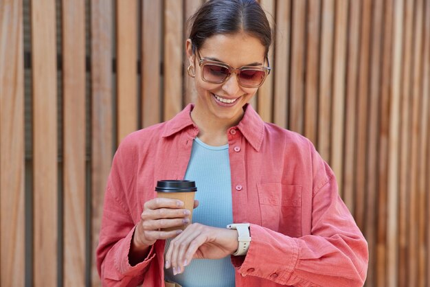 Positieve brunette vrouw drinkt afhaalmaaltijden cafeïne drank geniet van vrije tijd overdag kijkt naar smartwatch draagt een zonnebril en casual roze shirt vormt tegen houten vandaar Vrije tijd en levensstijl