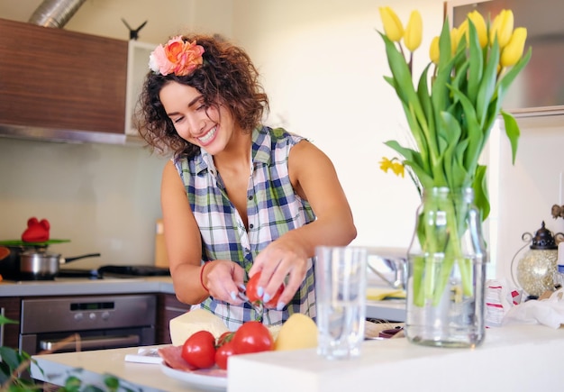 Positieve brunette vrouw met krullend haar maakt salade met tomaten en aardappel in een huiskeuken.