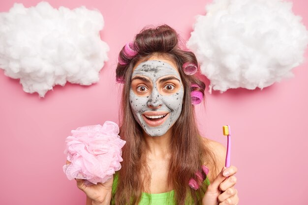 Positieve brunette vrouw past haarrollers toe voor het maken van kapsel houdt douchespons borstelt tanden met borstel heeft voedend kleimasker op gezicht geïsoleerd over roze muur