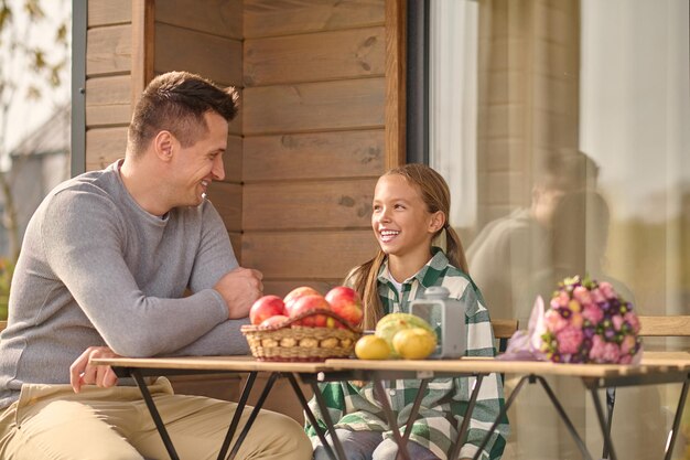 Positieve communicatie. Glimlachend jonge volwassen man in grijze trui en vrolijk schoolmeisje in geruite hemd kijken elkaar zittend aan tafel op veranda op mooie dag