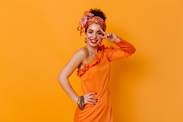 Positieve dame in oranje elegante jurk en modieuze hoofdband glimlacht oprecht en toont vredesteken op oranje ruimte.