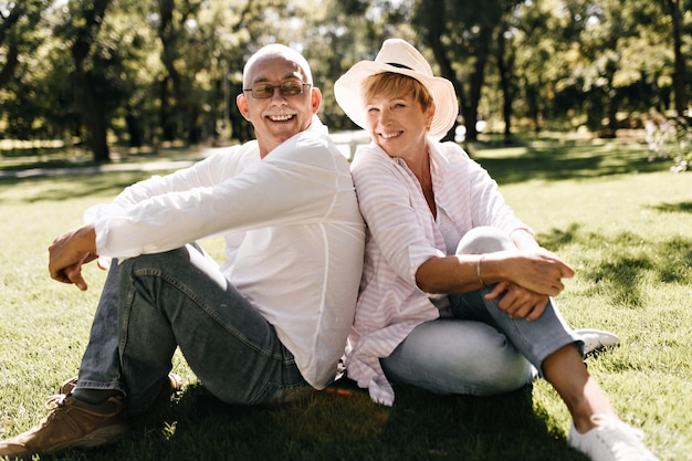 Positieve dame met coole hoed in gestreepte stijlvolle blouse en spijkerbroek glimlachend en zittend op het gras met man in brillen en licht shirt buiten.