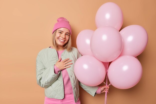 Positieve dankbare vrouw blij gefeliciteerd met verjaardag draagt hoed en jas poses met bos van helium ballonnen geïsoleerd over bruine achtergrond mensen emoties viering concept