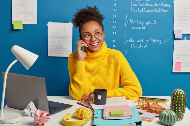 Positieve donkere huid meisje maakt telefoongesprek, bespreekt verbetering en ontwikkeling van zakelijk project, gekleed in gele trui, kijkt opzij, vormt tegen blauwe achtergrond