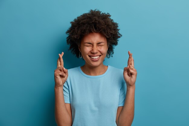 Positieve donkere vrouw met afro kapsel, kruist vingers voor geluk, sluit ogen en glimlacht breed, gelooft dat dromen uitkomen, draagt casual t-shirt, geïsoleerd op blauwe achtergrond