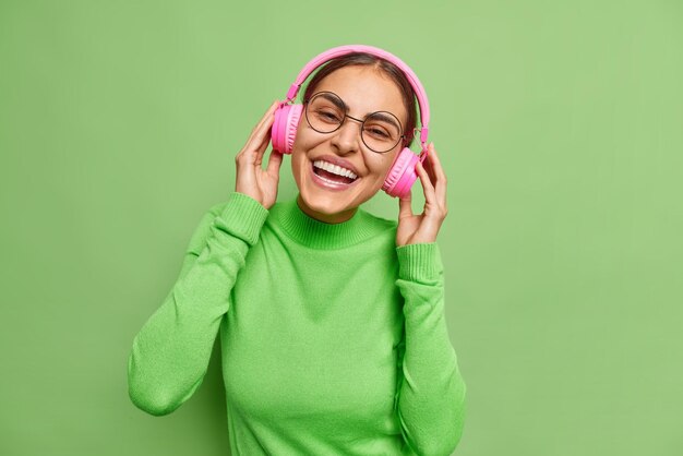 Positieve donkerharige Europese vrouw houdt handen op roze stereo hoofdtelefoon geniet van luisteren muziek glimlacht tandjes gekleed in casual coltrui geïsoleerd over groene achtergrond Monochroom schot