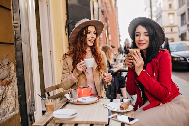 Positieve donkerharige vrouw in rode jas genieten van koffie met gesloten ogen met haar vriend