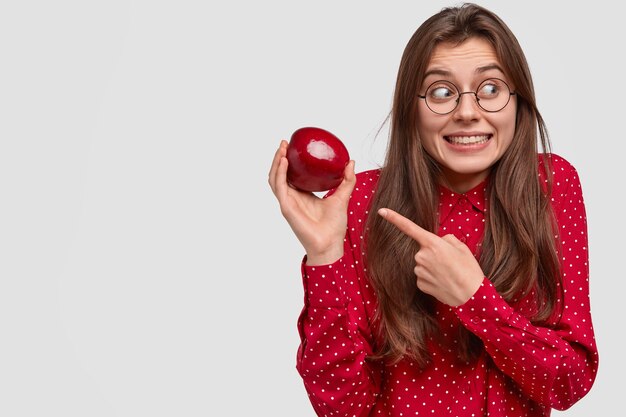 Positieve donkerharige vrouw met brede glimlach, wijst naar vers fruit, kijkt vrolijk opzij, gekleed in een rode blouse met stippen