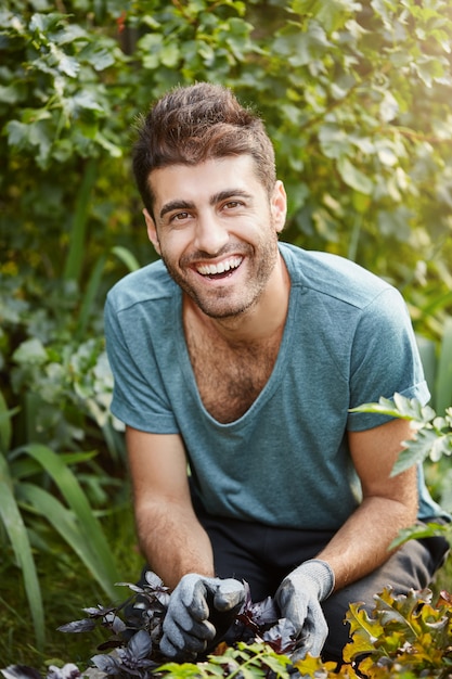 Positieve emoties, gezonde levensstijl. Verticale close-up buitenshuis portret van jonge volwassen bebaarde Kaukasische mannelijke tuinman in blauw t-shirt glimlachen, werken in de tuin,