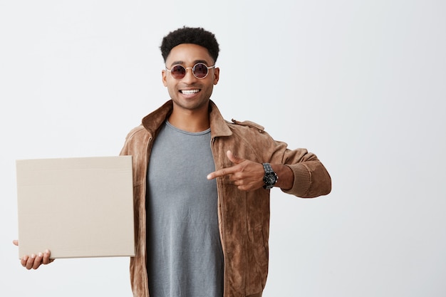 Positieve emoties. sluit omhoog van jonge knappe afro amerikaanse mannelijke student met krullend haar in vrijetijdskleding en zonnebril houdend kartonraad, wijzend op het met hand, glimlachend met tanden.