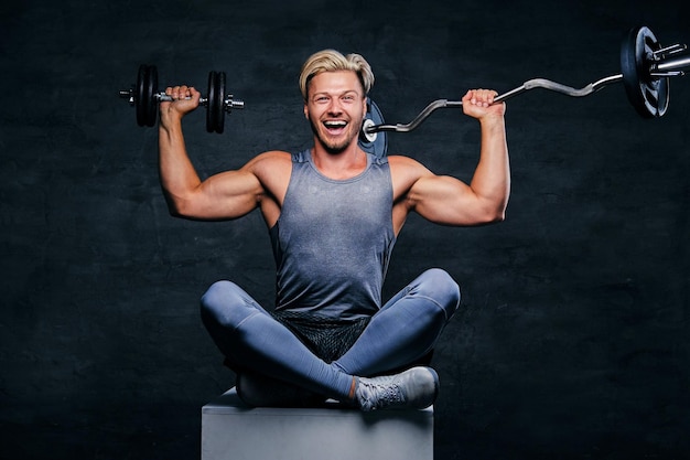 Positieve en lachende blonde sportieve man is gekleed in een grijze sportkleding zit op een doos en houdt barbeel vast.