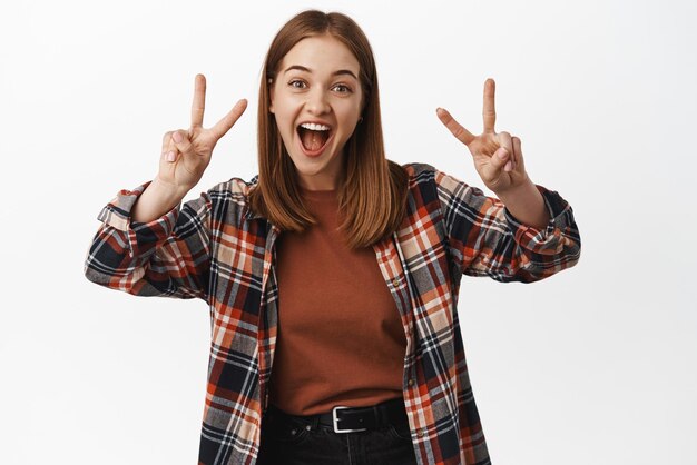 Positieve en vriendelijke jonge vrouw toont vrede vsign gebaar lachen en glimlachen poseren gelukkig op camera staande in vrijetijdskleding tegen witte achtergrond