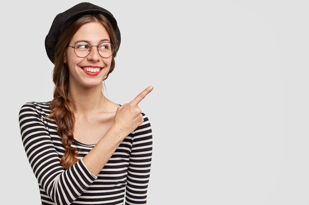 Positieve Franse vrouw met aantrekkelijke uitstraling, vrolijke uitdrukking, wijst met wijsvinger opzij, toont lege ruimte, gekleed in elegante kleding