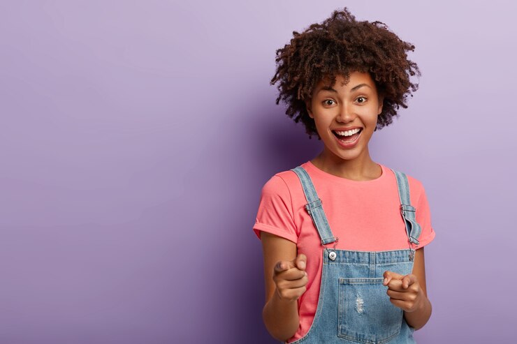Gratis Foto positieve gekrulde jongedame selecteert je, wijst met beide wijsvingers naar de camera, lacht vrolijk, draagt vrijetijdskleding, is blij en tevreden, staat tegen een paarse achtergrond. jij bent mijn type