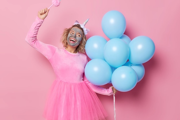 Positieve glimlachende zorgeloze vrouw bereidt zich voor op vakantie past klei voedend masker op gezicht vast houdt bos blauwe heliumballonnen draagt feestelijke jurk danst zorgeloos geïsoleerd over roze achtergrond