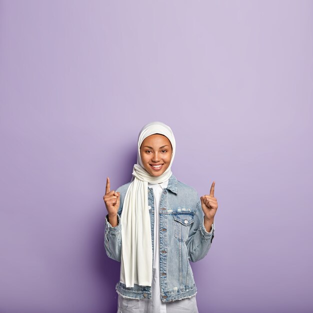 Positieve, goed uitziende moslimvrouw wijst naar boven, reageert op geweldige promo-kopie ruimte