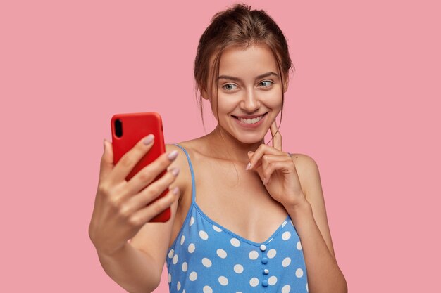 Positieve jonge blanke vrouw neemt foto van zichzelf met moderne mobiele telefoon, heeft een tedere glimlach op het gezicht, draagt blauwe polka dor jurk, modellen tegen roze muur. Mooie dame poseert voor selfie