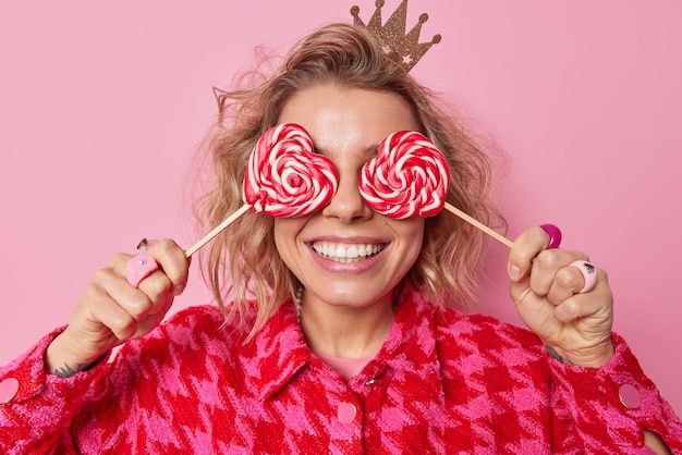 Positieve jonge blonde vrouw bedekt ogen met zoete snoepjes op stokken glimlacht breed heeft witte tanden draagt een kleine kroon op het hoofd en geruite jas heeft een vrolijke stemming geïsoleerd over roze achtergrond