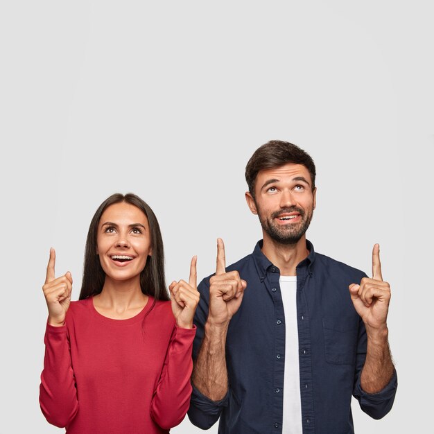 Gratis foto positieve jonge europese brunette schattige vrouw met gebruinde huid, staan in de buurt van haar minnaar, wijs samen naar boven