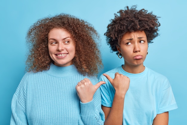 Positieve jonge Europese vrouw met krullend borstelig haar en droevige, donkere vrouwelijke duimen naar elkaar wijzen erop dat ze ervoor moeten kiezen om dicht bij elkaar in blauwe kleding te gaan staan. Zij is schuldig, niet ik