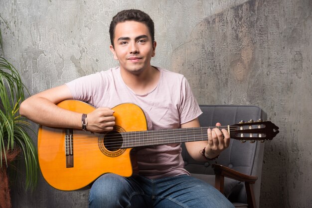 Positieve jonge man zittend op een stoel en gitaar spelen.