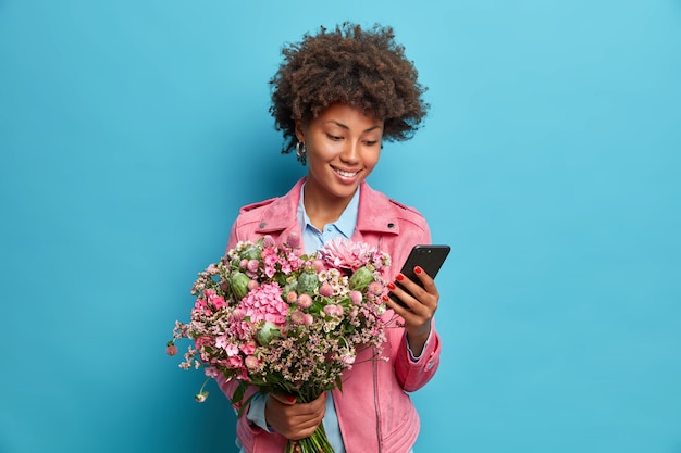 Gratis foto positieve jonge vrouw accepteert gefeliciteerd met haar verjaardag houdt mobiele telefoon boeket bloemen draagt roze jas geïsoleerd over blauwe muur controleert newsfeed