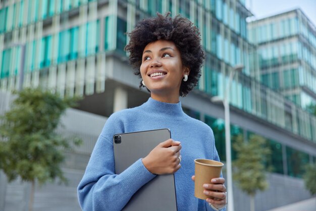 Positieve jonge vrouw houdt digitale tablet gebruikt moderne gadget drankjes afhaalmaaltijden koffie glimlacht vrolijk draagt casual blauwe trui poses in stedelijke omgeving komt terug van studeren. Mensen en levensstijl concept