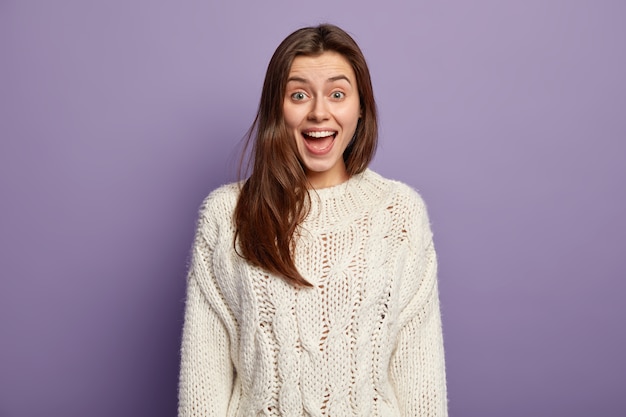 Positieve jonge vrouw met blije uitdrukking, opent mond van verbazing, reageert op positief onverwacht nieuws, draagt witte trui, staat tegen violette muur, geïntrigeerd door iets te vertellen