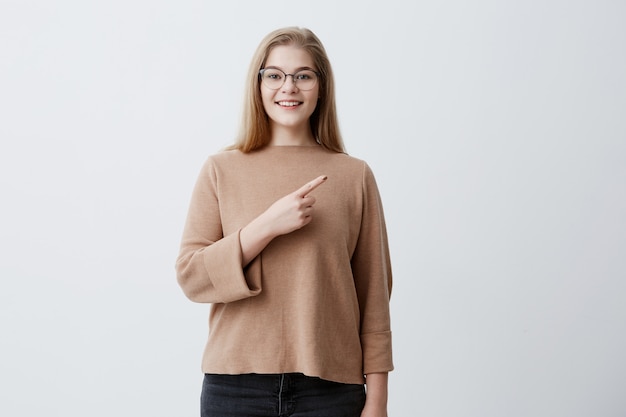 Positieve jonge vrouw met blond haar, bruine trui en bril dragen, heeft een gezonde huid, aangename glimlach, wijst op kopie ruimte op een grijze achtergrond. Moet je zien! Reclame concept