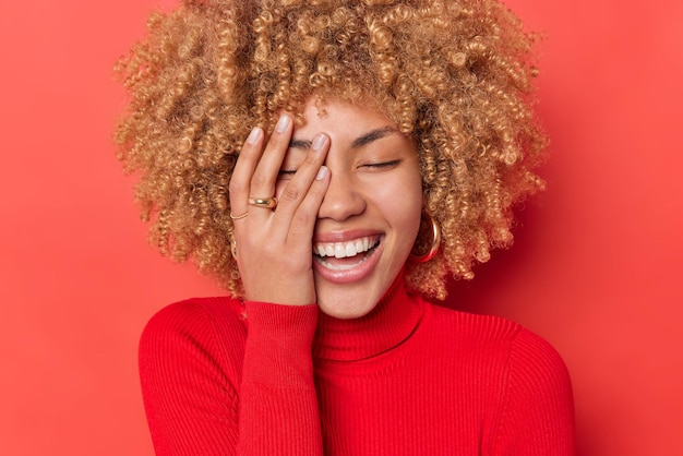 Positieve jonge vrouw met krullend blond haar giechelt vrolijk en maakt gezichtpalm oprechte emoties uitdrukt en draagt een casual coltrui geïsoleerd op rode achtergrond. Mensen geluk en vreugde concept.