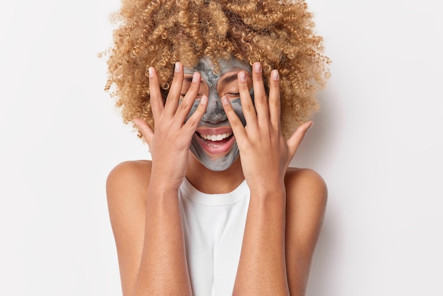 Positieve jonge vrouw met krullend borstelig haar houdt handen over het gezicht en probeert het kleimasker van het gezicht te verbergen voor huidverzorging heeft een vrolijke stemming geïsoleerd op een witte achtergrond. schoonheid en wellness-concept.
