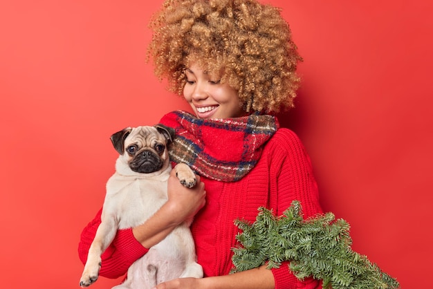 Positieve jonge vrouw met krullend haar houdt haar rashond vast van de kerstmarkt en draagt een groene sparrenkrans gekleed in winterkleren geïsoleerd op rode achtergrond. vakantie voorbereiding