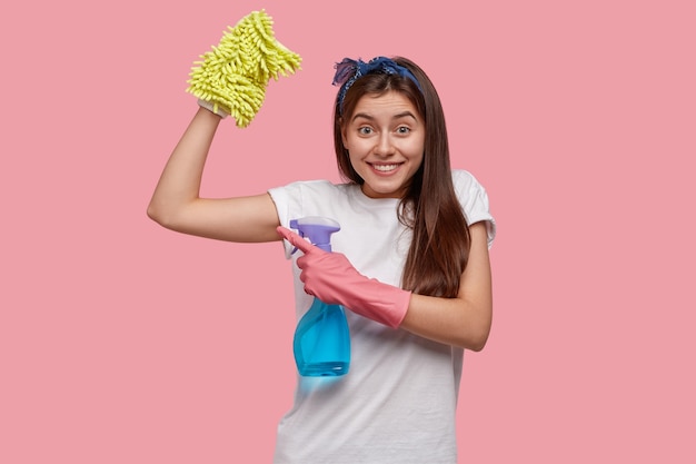 Positieve jonge vrouw toont spieren na moe werk over huis, gekleed in wit casual t-shirt, houdt een fles spray