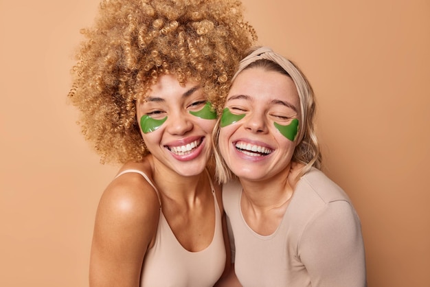 Positieve jonge vrouwelijke vrienden glimlachen in het algemeen veel plezier hebben samen groene hydrogelpleisters toepassen onder de ogen ondergaan schoonheidsprocedures terloops gekleed poseren tegen beige achtergrond Huidverzorgingsconcept