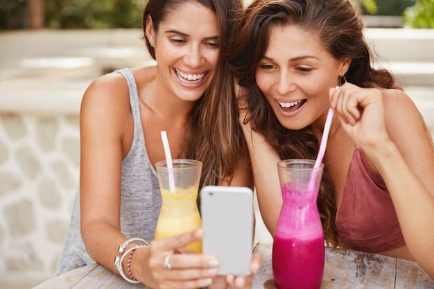Positieve jonge vrouwtjes met gelukkige vrouwtjes kijken naar grappige video's op de smartphone, drinken cocktails