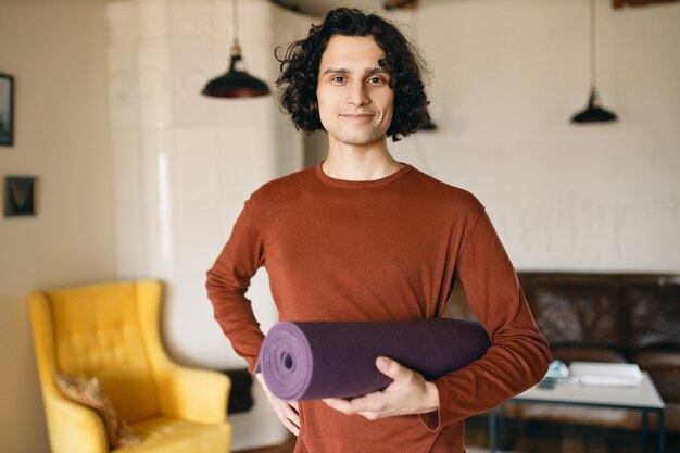 Positieve jongeman in vrijetijdskleding met rol yogamat onder zijn arm gaat binnenshuis oefenen, blij om thuis te blijven tijdens quarantaine, meer tijd te besteden aan zelfontwikkeling en gezonde activiteit