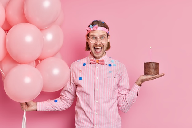 Positieve knappe Europese man draagt shirt met strik houdt kleine chocoladetaart vast en stelletje opgeblazen ballonnen geniet van verjaardagsfeestje geïsoleerd over roze muur