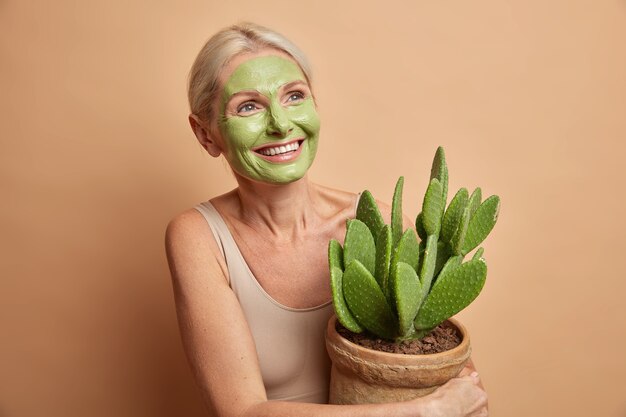 Positieve knappe Europese vrouw senior vrouw past groene schoonheid masker houdt cactus in pot gekleed in vrijetijdskleding geïsoleerd over beige muur