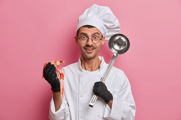 Positieve kok poseert met ongekookte rivierkreeftjes, stalen pollepel, gaat smakelijke soep bereiden, draagt een wit uniform