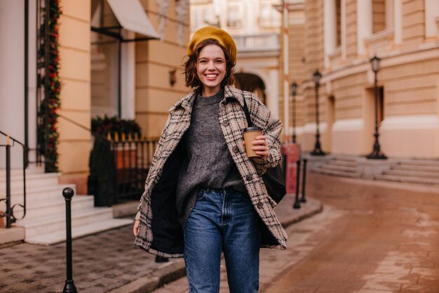 Positieve kortharige vrouw in stijlvolle flodderige kleding lacht Portret van een dame met een kopje thee op de achtergrond van prachtige gebouwen