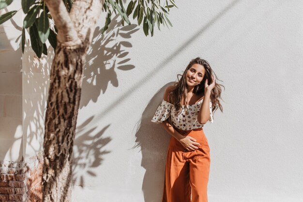 Positieve langharige dame in oranje zomerbroek met glimlach kijkt naar voren op witte muur met olijfboom