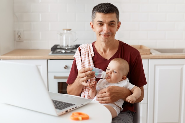 Positieve man met een glimlach die een bordeauxrood casual t-shirt draagt met een handdoek op zijn schouder, voor de baby zorgt en online vanuit huis werkt, zijn dochter water geeft, kijkt naar de camera.