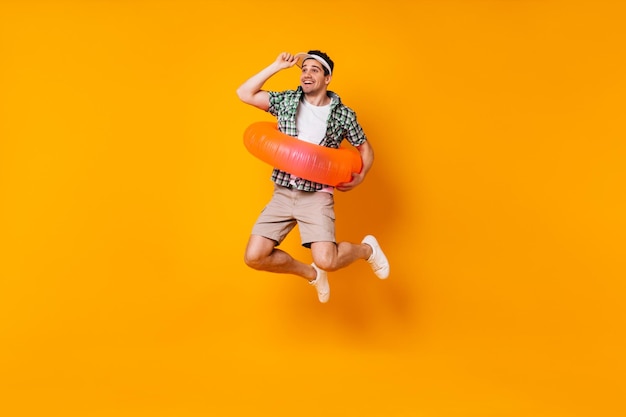 Positieve man springt op oranje achtergrond Portret van man in pet en zomeroutfit met opblaasbare cirkel