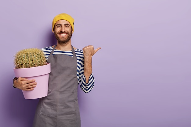 Positieve mannelijke tuinman wijst met duim weg, toont lege ruimte voor uw advertentie, houdt pot met cactusachtergrond vast