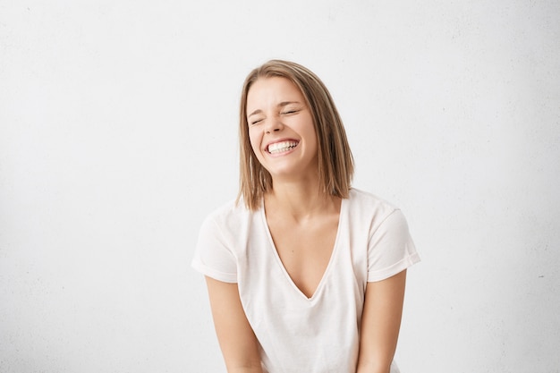 Positieve menselijke emoties. Headshot van een gelukkig emotionele tienermeisje met bob-kapsel lachend uit de grond van haar hart, de ogen gesloten houden, perfecte witte tanden laten zien terwijl ze binnenshuis plezier heeft