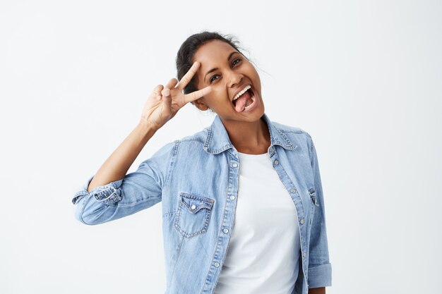 Positieve menselijke emoties. portret van ontspannen zorgeloze Afro-Amerikaanse vrouw met een glimlach die haar tong toont en een V-teken draagt een spijkerblouse die één oog strak sluit terwijl ze plezier heeft met vrienden binnen